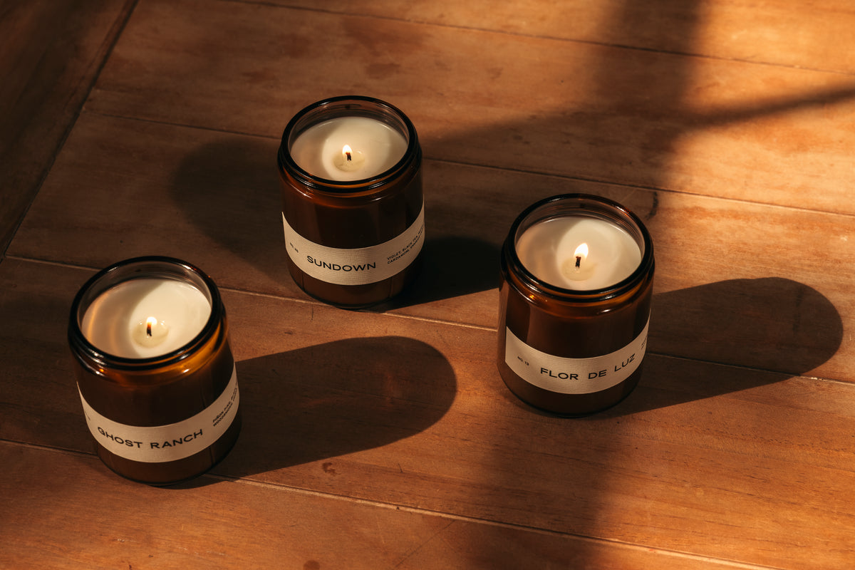 Three lit amber glass jar candles with white labels are arranged on a wooden floor in warm, dramatic sunlight and shadow.